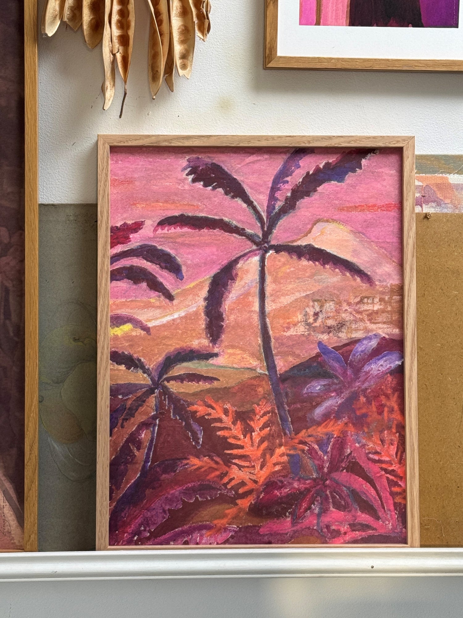 Framed painting of palm trees and mountains in warm pink, purple, and orange tones on white shelf with dried plants above