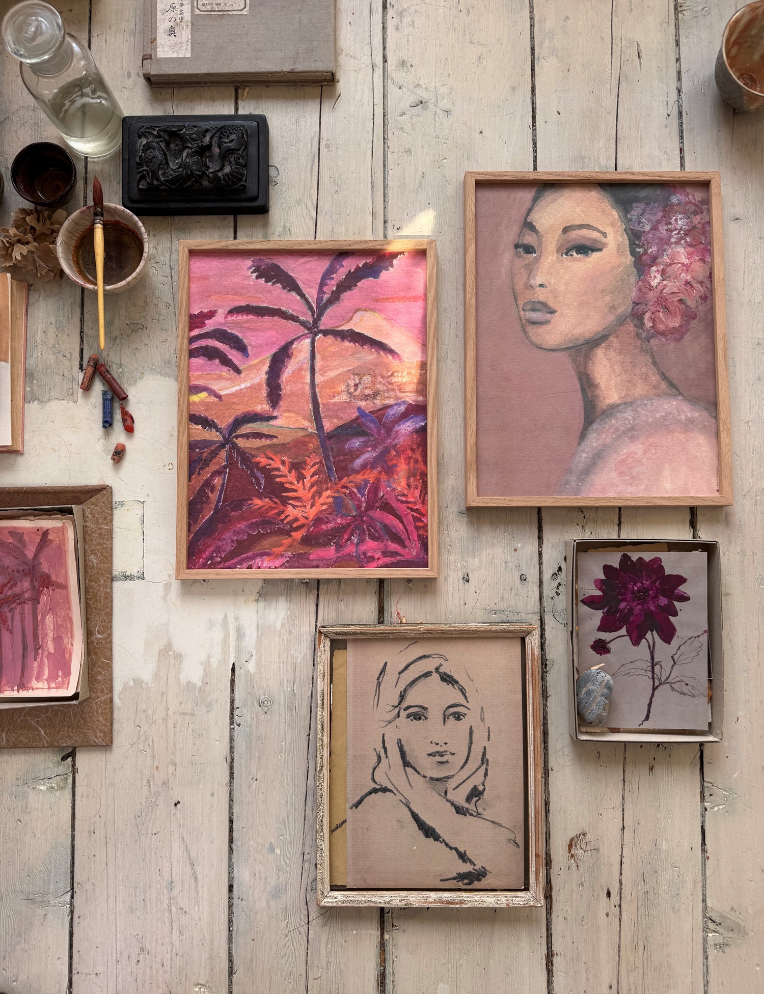 Framed tropical painting, woman’s portrait, small sketch, pressed flower, and stone arranged on a worn wooden floor