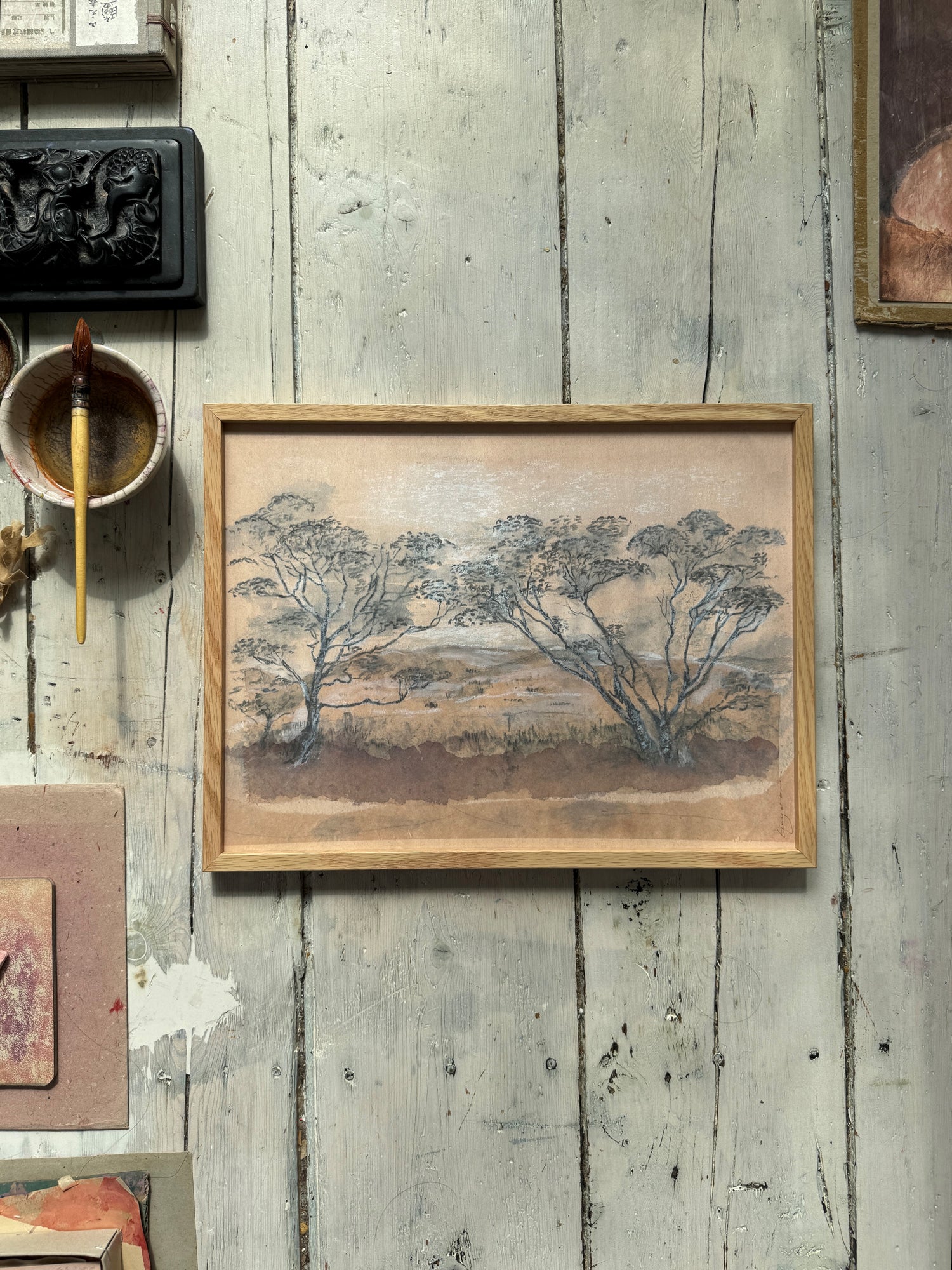 Framed landscape painting with trees and brownish tones on rustic wooden floor with paintbrush and bowls nearby.
