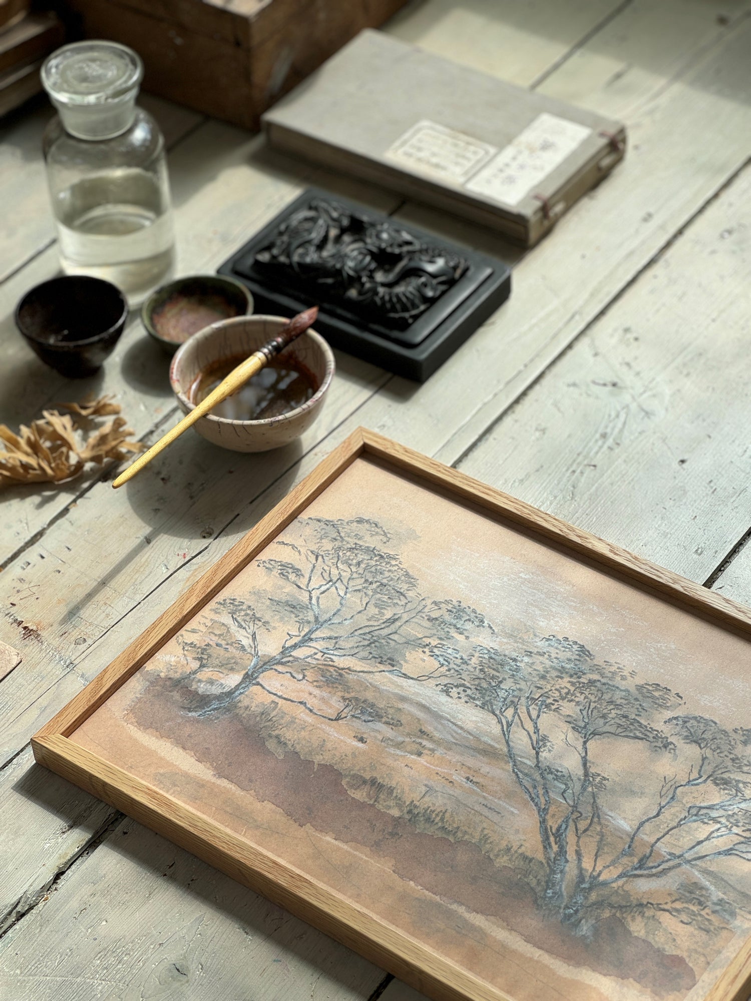 Close-up of framed tree landscape painting on wooden floor with art supplies and a black carved box.
