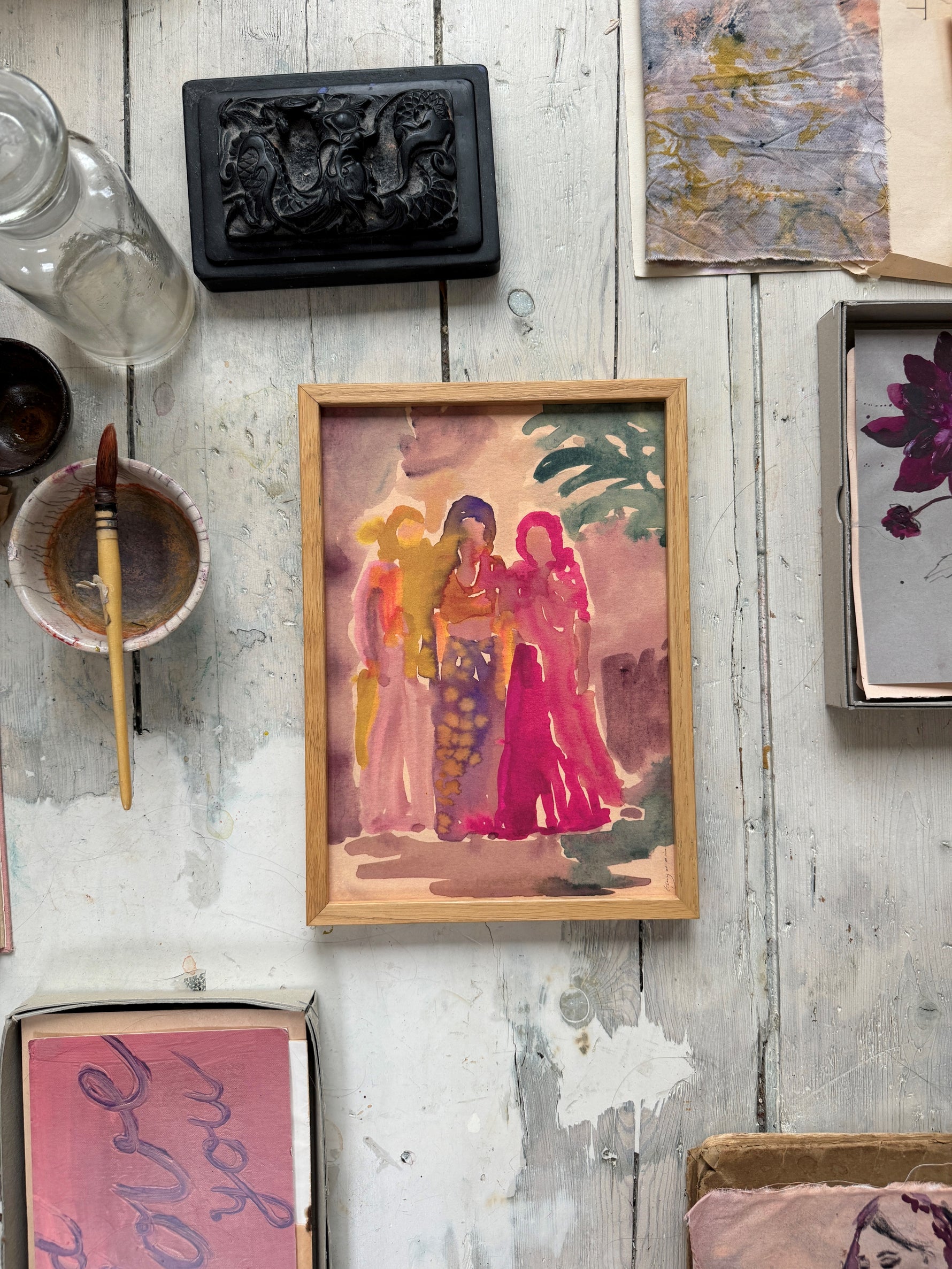 Framed watercolor painting of three women in colorful dresses with green leaves in background on rustic white wooden floor
