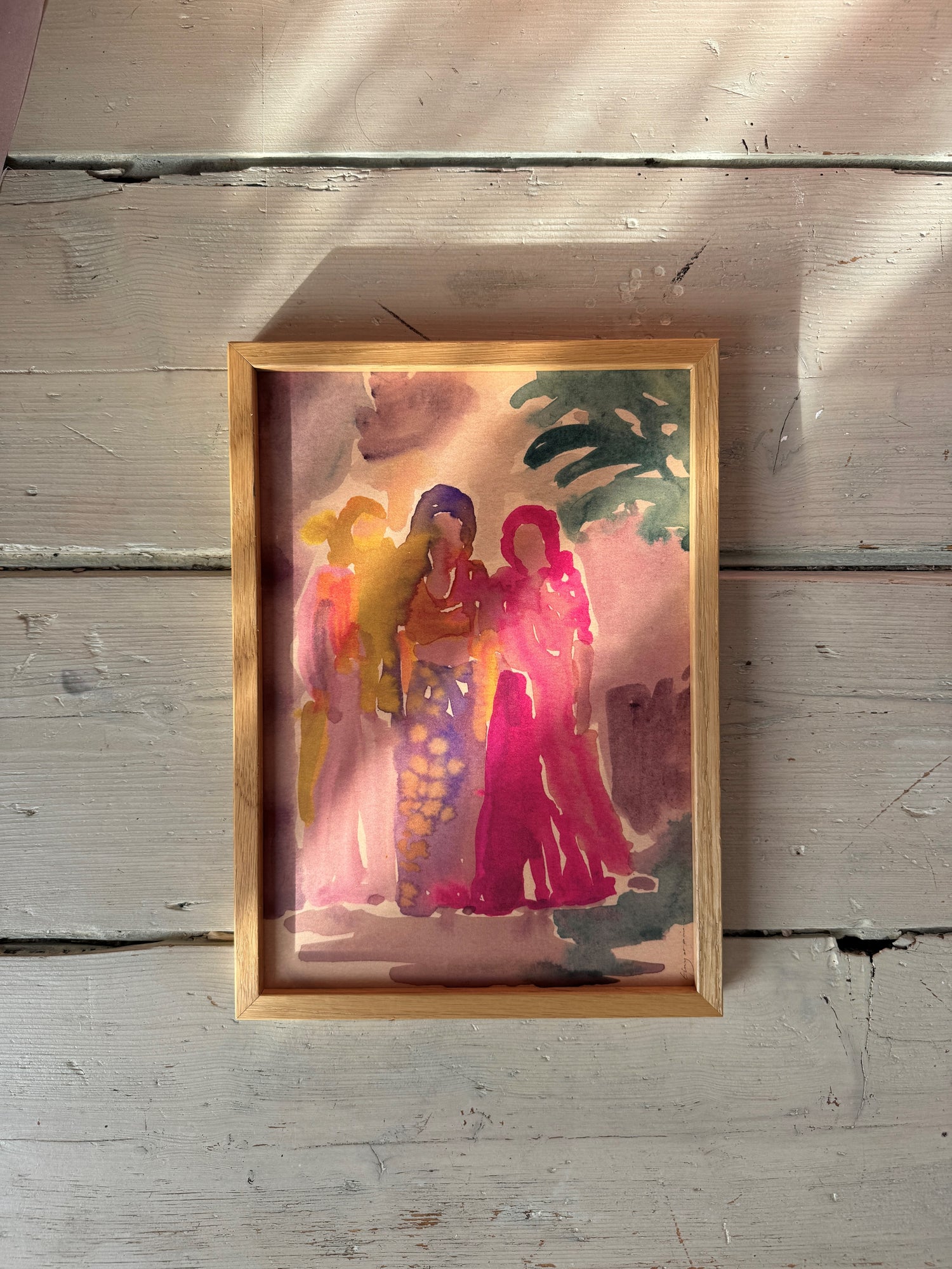 Top view of framed watercolor painting showing three women in vibrant dresses on weathered white wooden floor