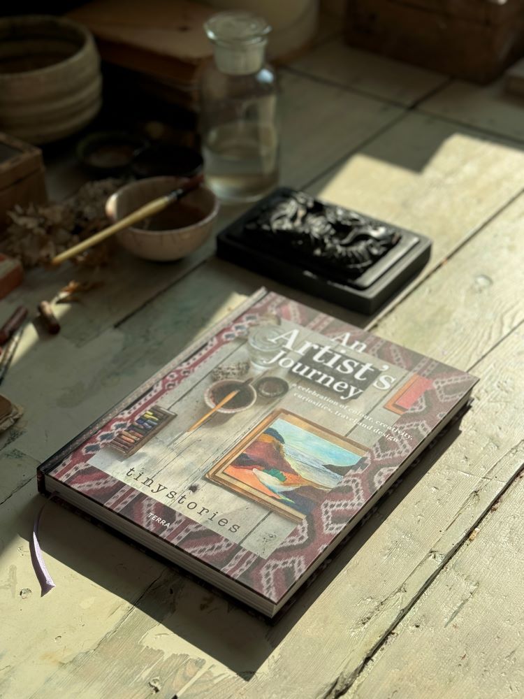 Hardcover book "An Artist's Journey" by tinystories on rustic wooden floor with art supplies and carved black box.