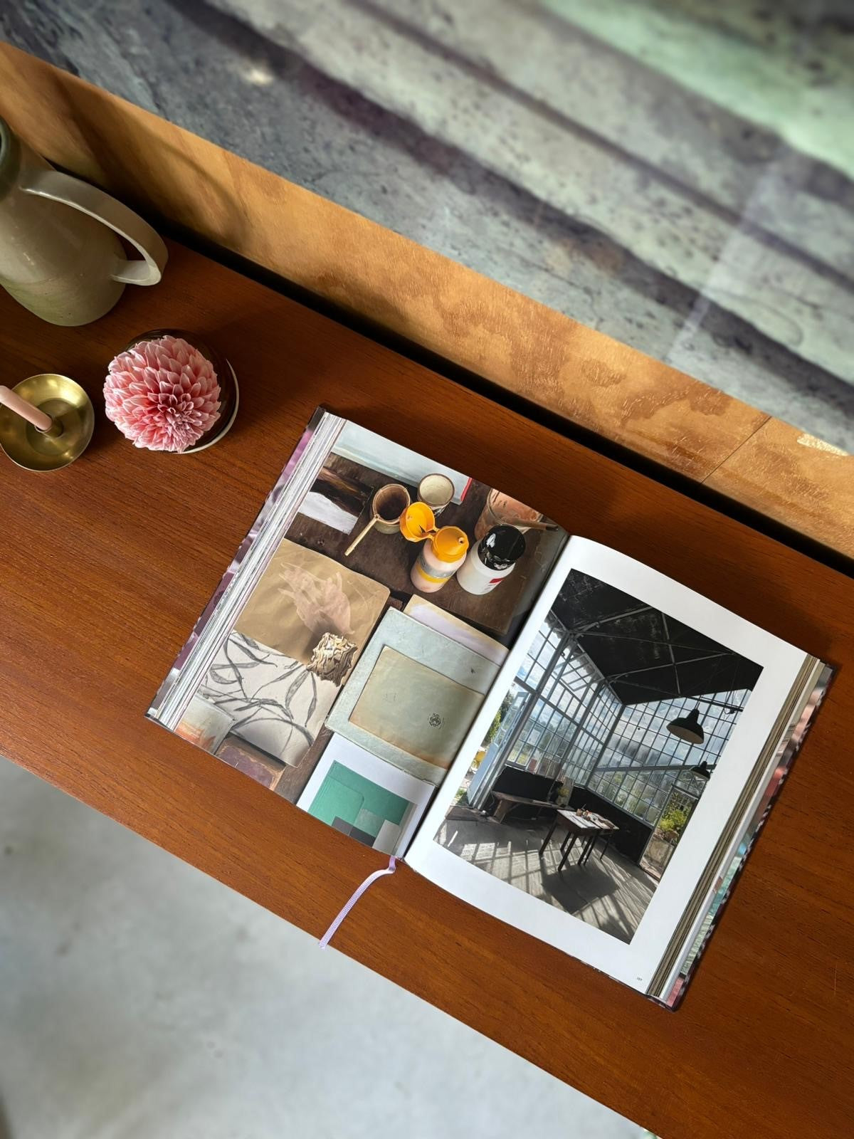Overhead view of open book on wooden desk showing art supplies and photo of sunlit interior with large windows.