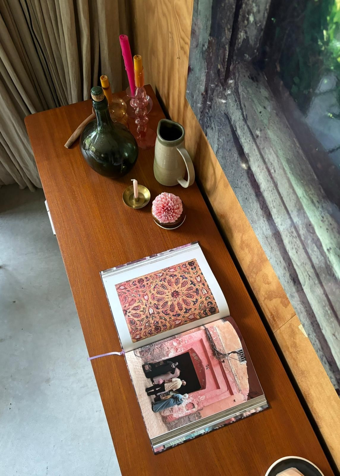 Overhead view of open book with detailed textile photo and pink building, on wooden table with vase and candle.