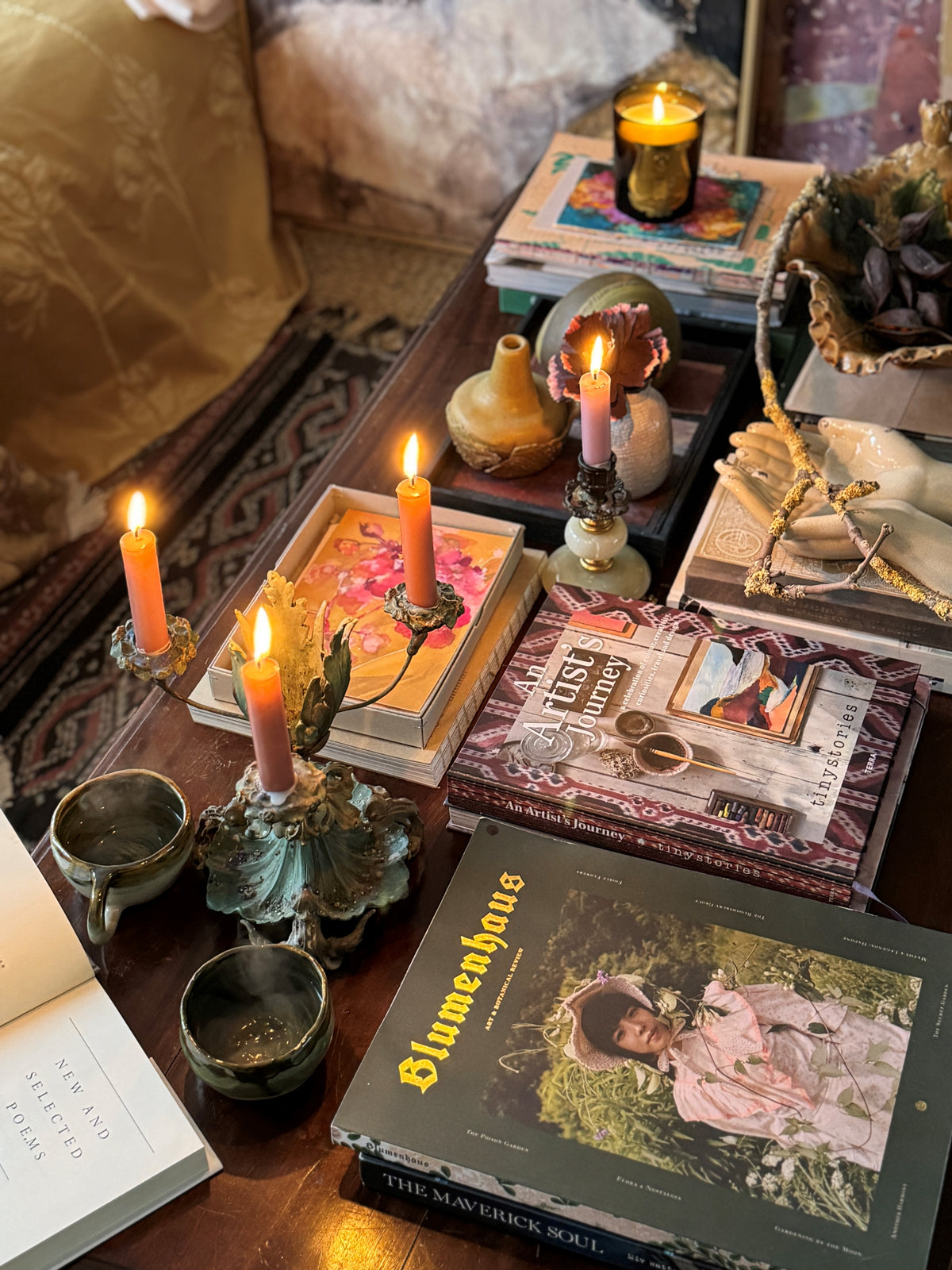 Cozy table setup with lit candles, books including "An Artist's Journey" and "Blumenhaus," and decorative ceramics.