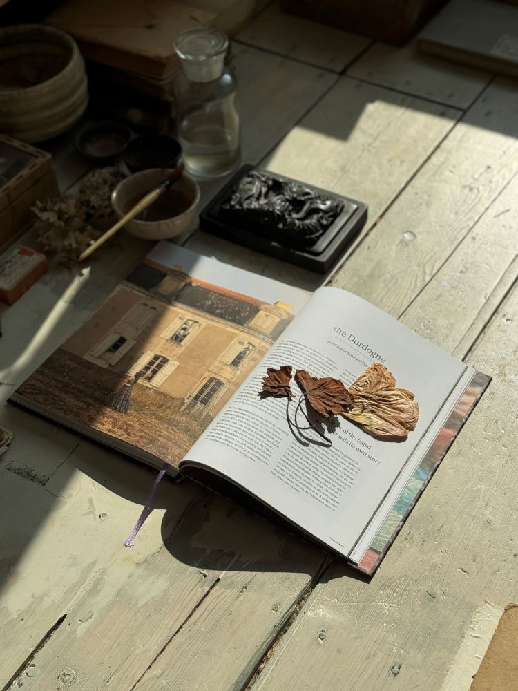 Open book with dried leaves on page titled "the Dordogne," lying on rustic wooden floor with art tools nearby.