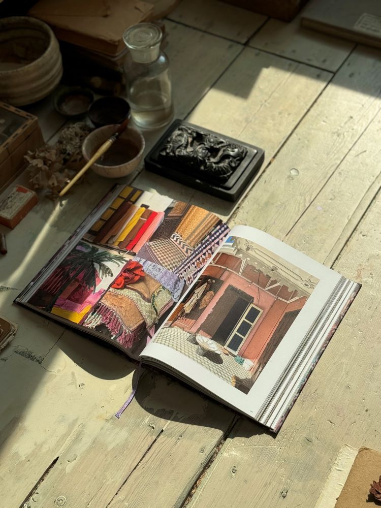 Open book on wooden floor showing colorful textiles and pink building under natural light.