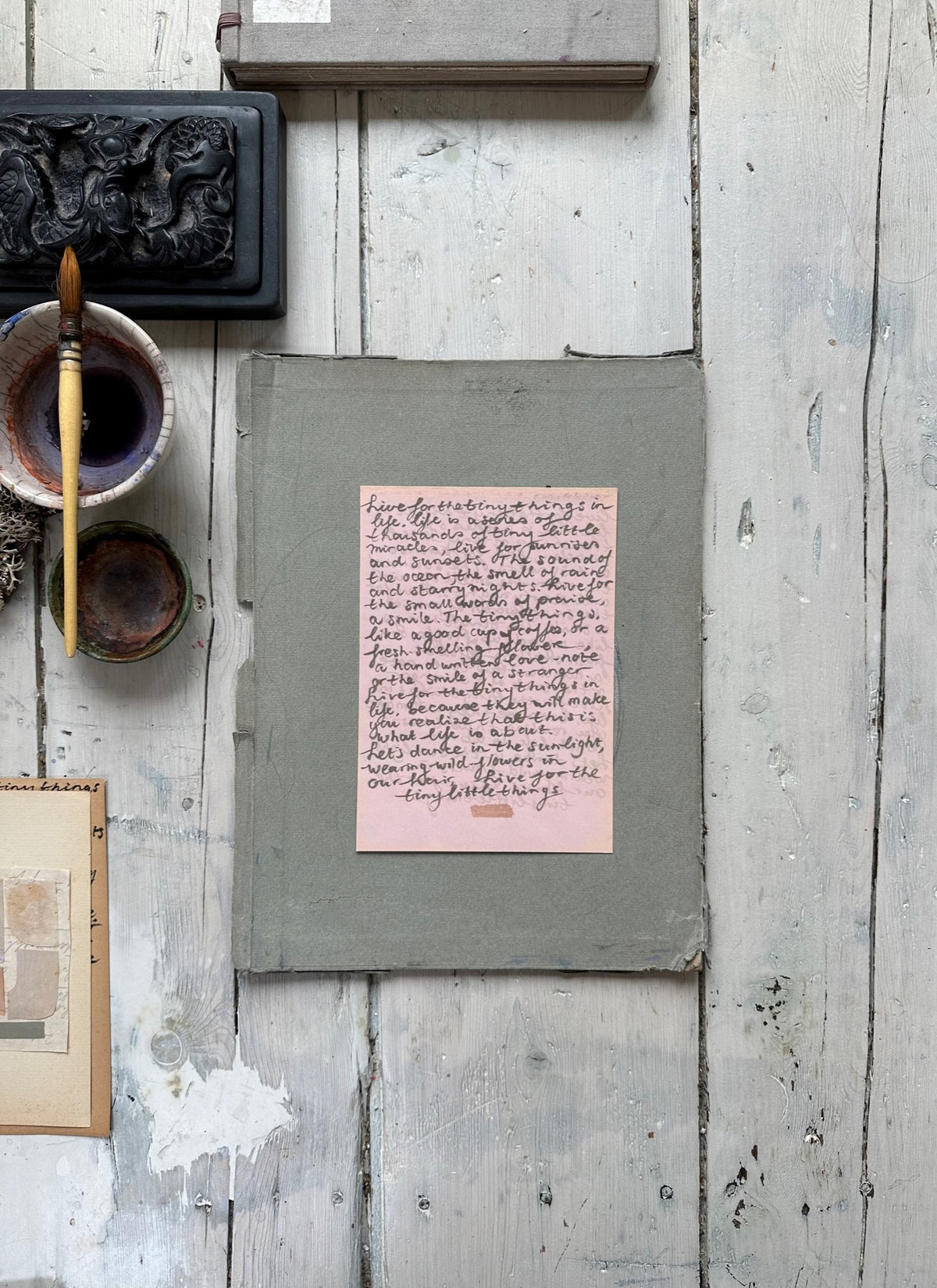 Pink handwritten note with cursive text on a small sheet inside a cardboard box with rustic wooden background.