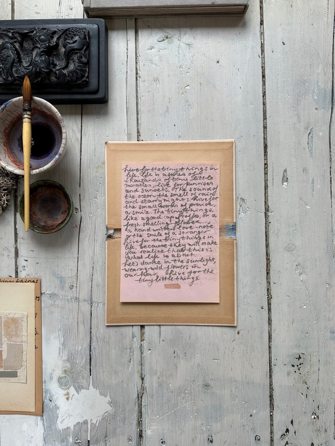 Pink handwritten note with cursive text on a small sheet inside a cardboard box with rustic wooden background.