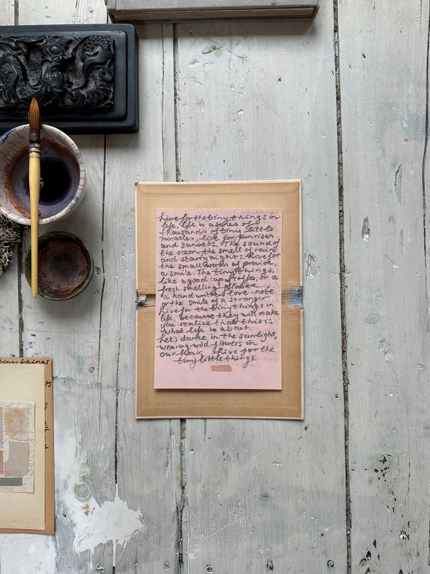 Pink handwritten note with cursive text on a small sheet inside a cardboard box with rustic wooden background.