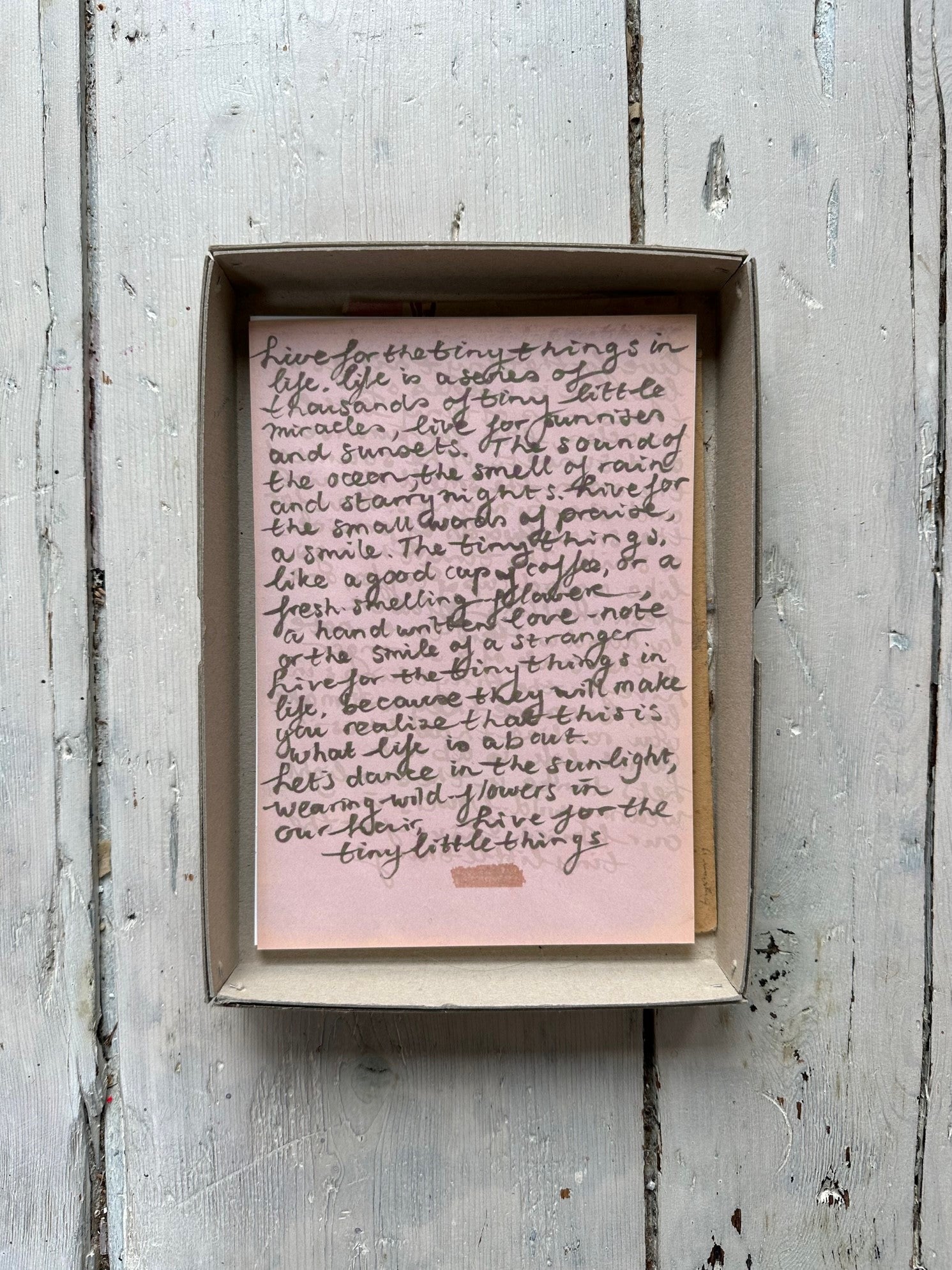 Pink handwritten note with cursive text on a small sheet inside a cardboard box with rustic wooden background.