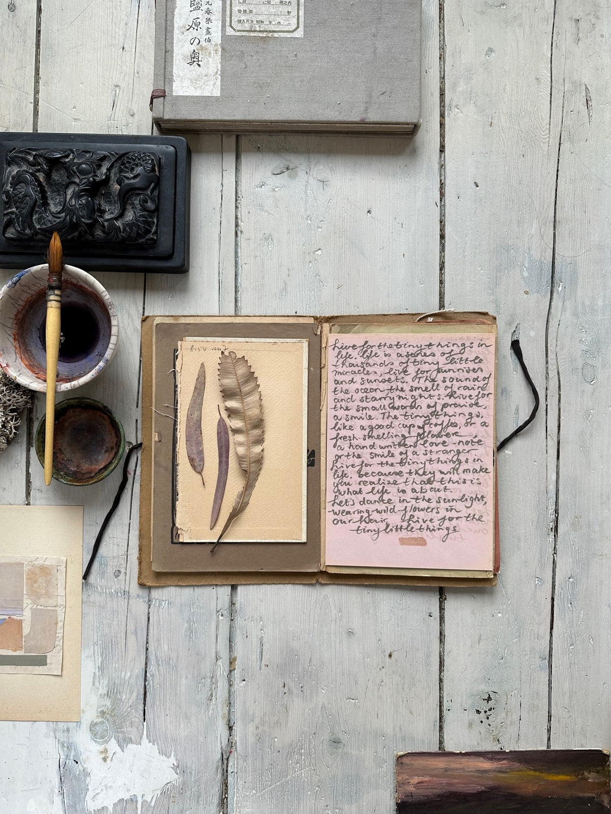 Open sketchbook on rustic wooden floor with three pressed leaves on the left page and a handwritten note on pink paper on the right page beside a black ink pot and brush.
