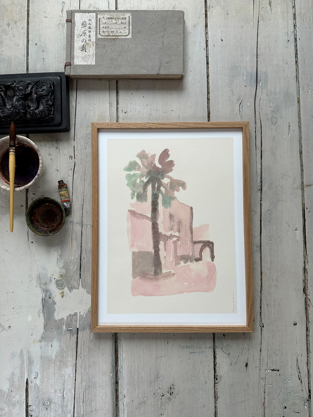 Framed watercolor painting of a palm tree and building in soft pink and green tones on rustic wooden floor with art supplies