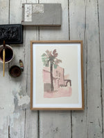 Framed watercolor painting of a palm tree and building in soft pink and green tones on rustic wooden floor with art supplies