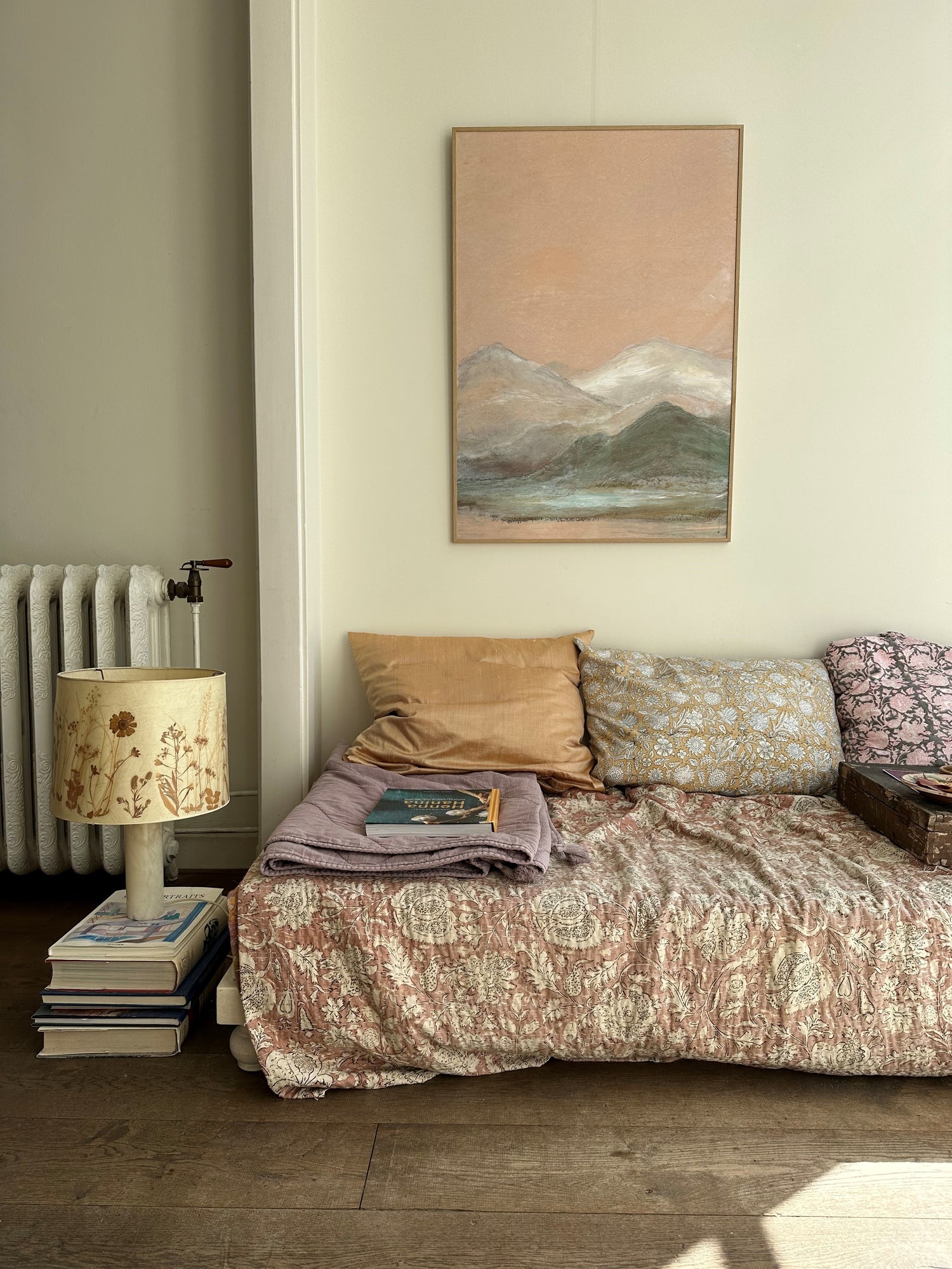 Cozy living room corner with framed mountain landscape painting, vintage lamp on stacked books, and patterned sofa