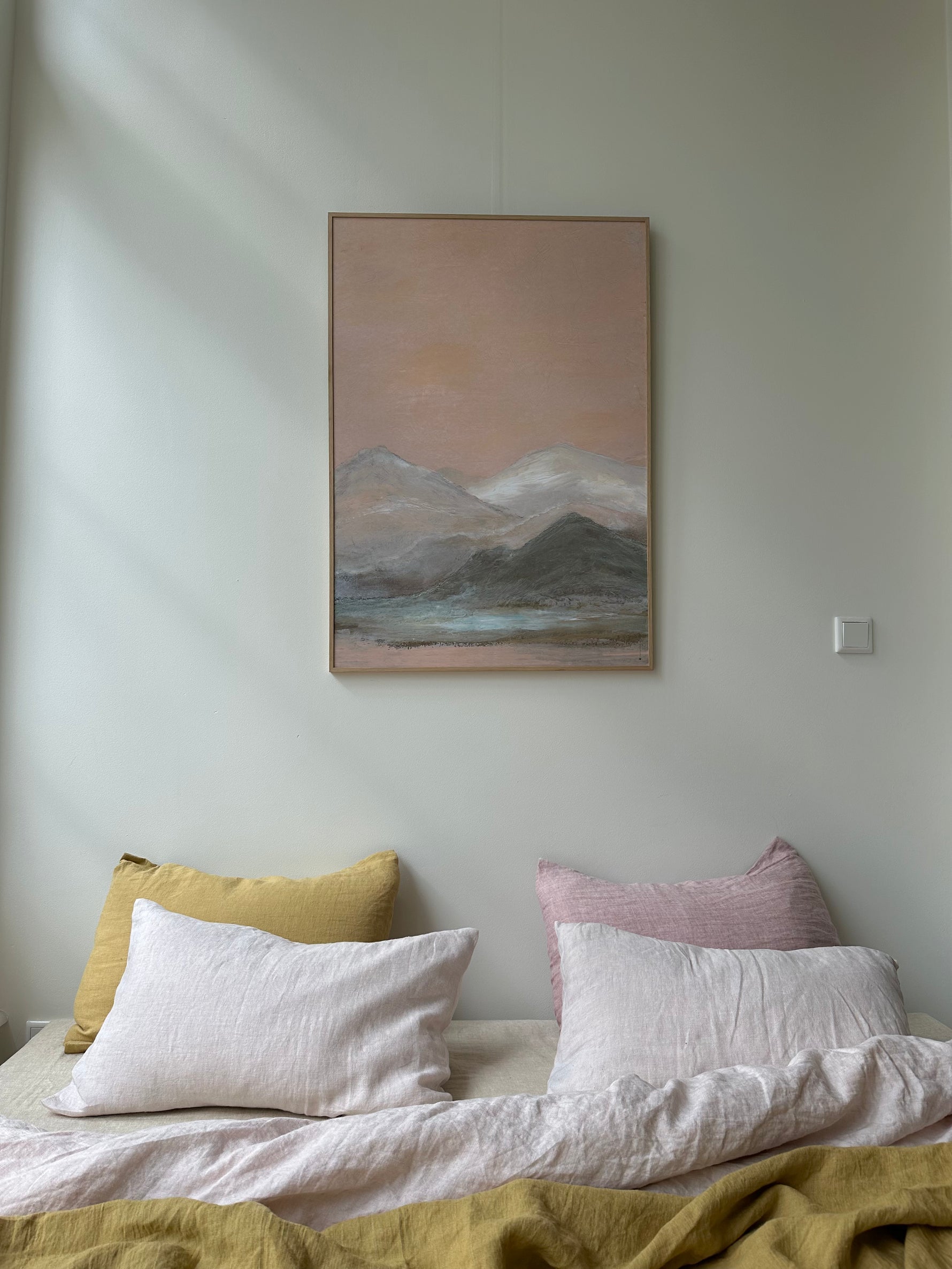 Framed soft mountain landscape painting with muted tones above bed with pastel cushions in bright bedroom