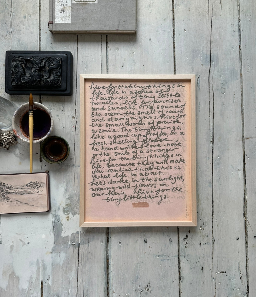 Framed handwritten poetic text artwork on soft pink background displayed on rustic wooden floor