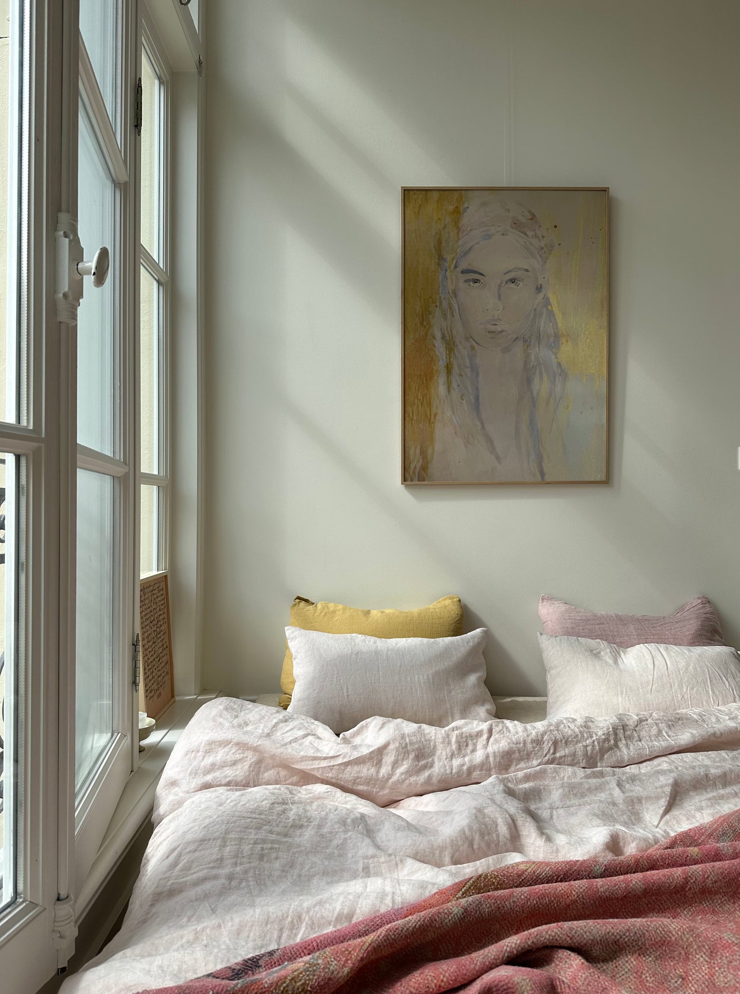 Soft pastel portrait painting of a woman hanging above cozy bed with textured pillows in bright bedroom