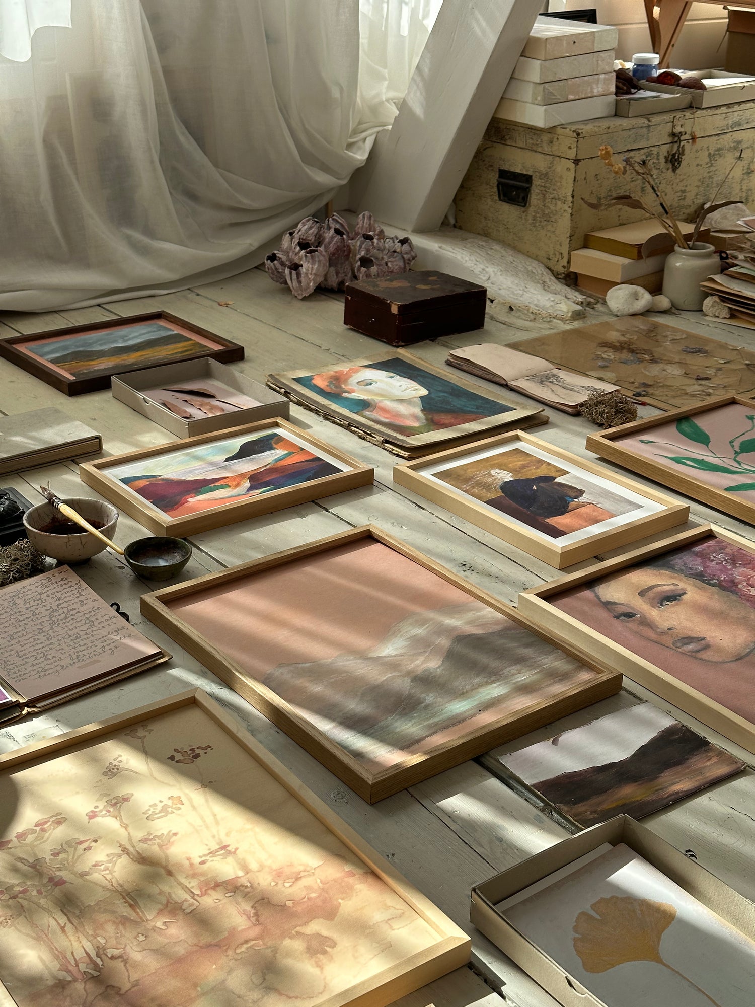 Collection of framed abstract and figurative artworks including mountain landscape laid out on rustic wooden floor with sunlight