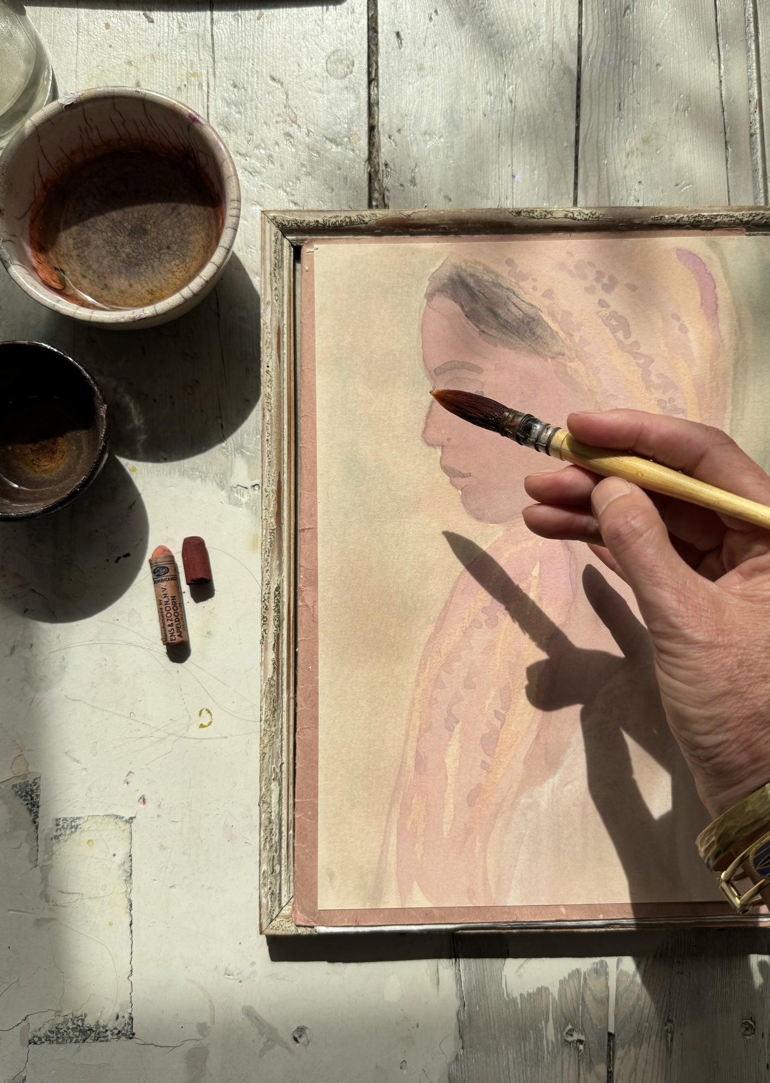 Close-up of hand holding paintbrush over framed watercolor portrait of woman in pink headscarf on wooden table with art tools