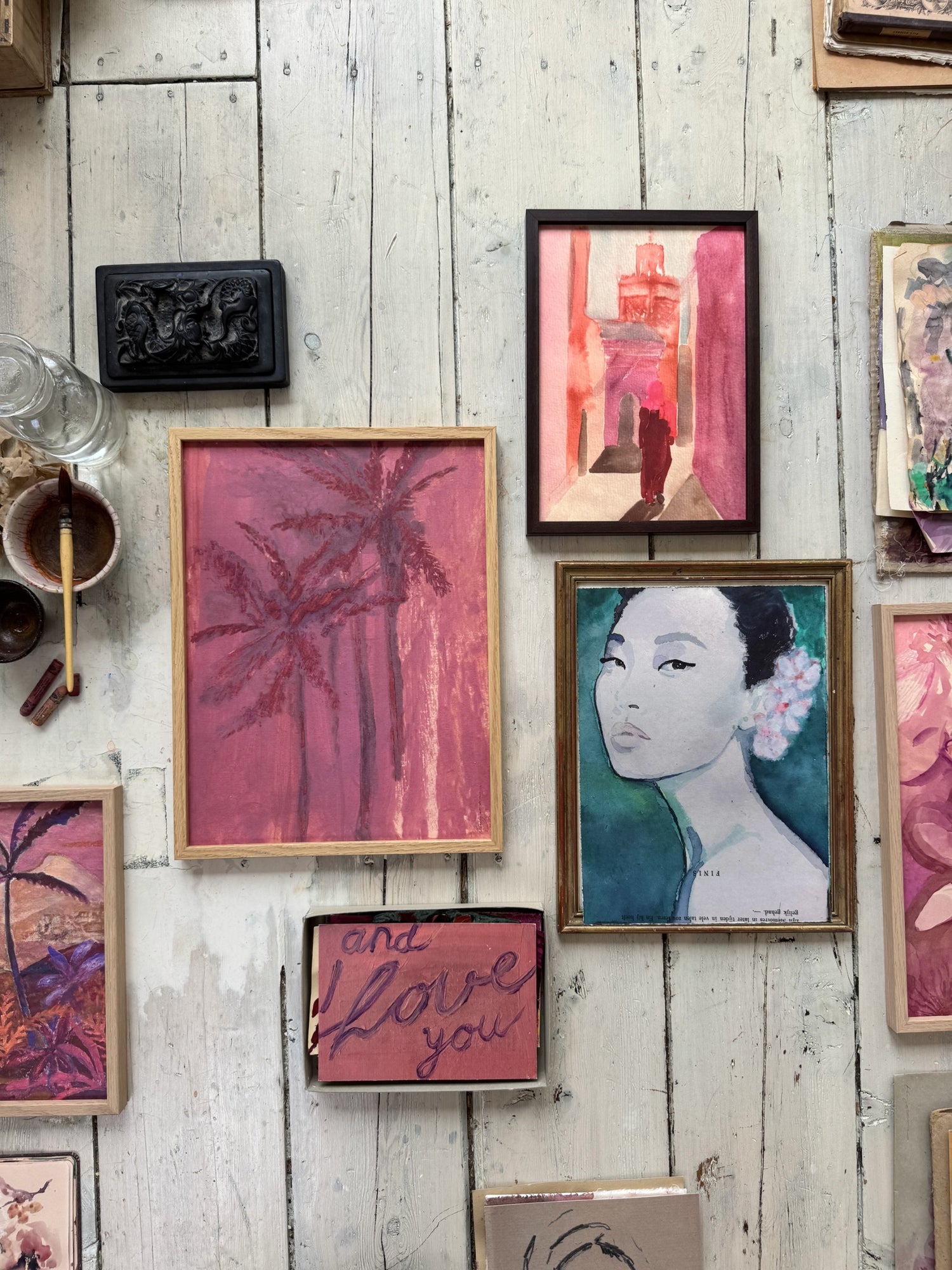 Floor display of multiple framed artworks including pink palm trees, woman portrait, floral paintings, and dried flowers on white wall