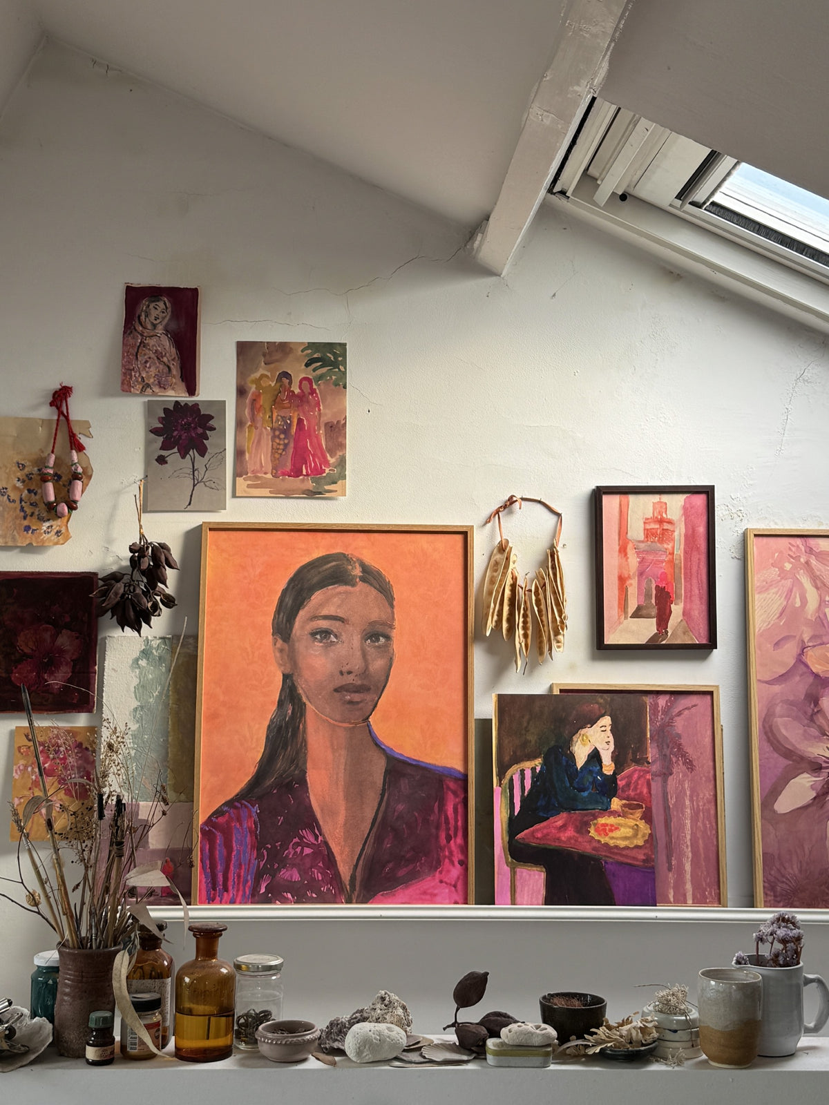 Wall display with framed portrait of woman, dried flowers, smaller paintings, and glass jars on white shelf in sunlit room