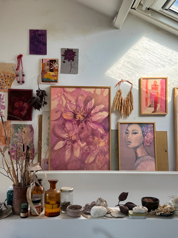Wall display with framed floral painting, woman portrait, dried flowers, and small art pieces above white shelf with jars

