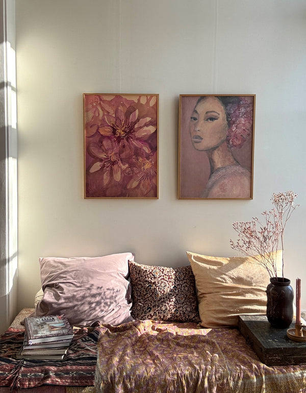 Close-up of two framed paintings of purple flowers and woman on white wall above colorful cushions on sofa