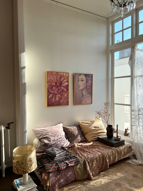 Cozy sunlit room with two framed paintings of flowers and woman on white wall above sofa with cushions and books