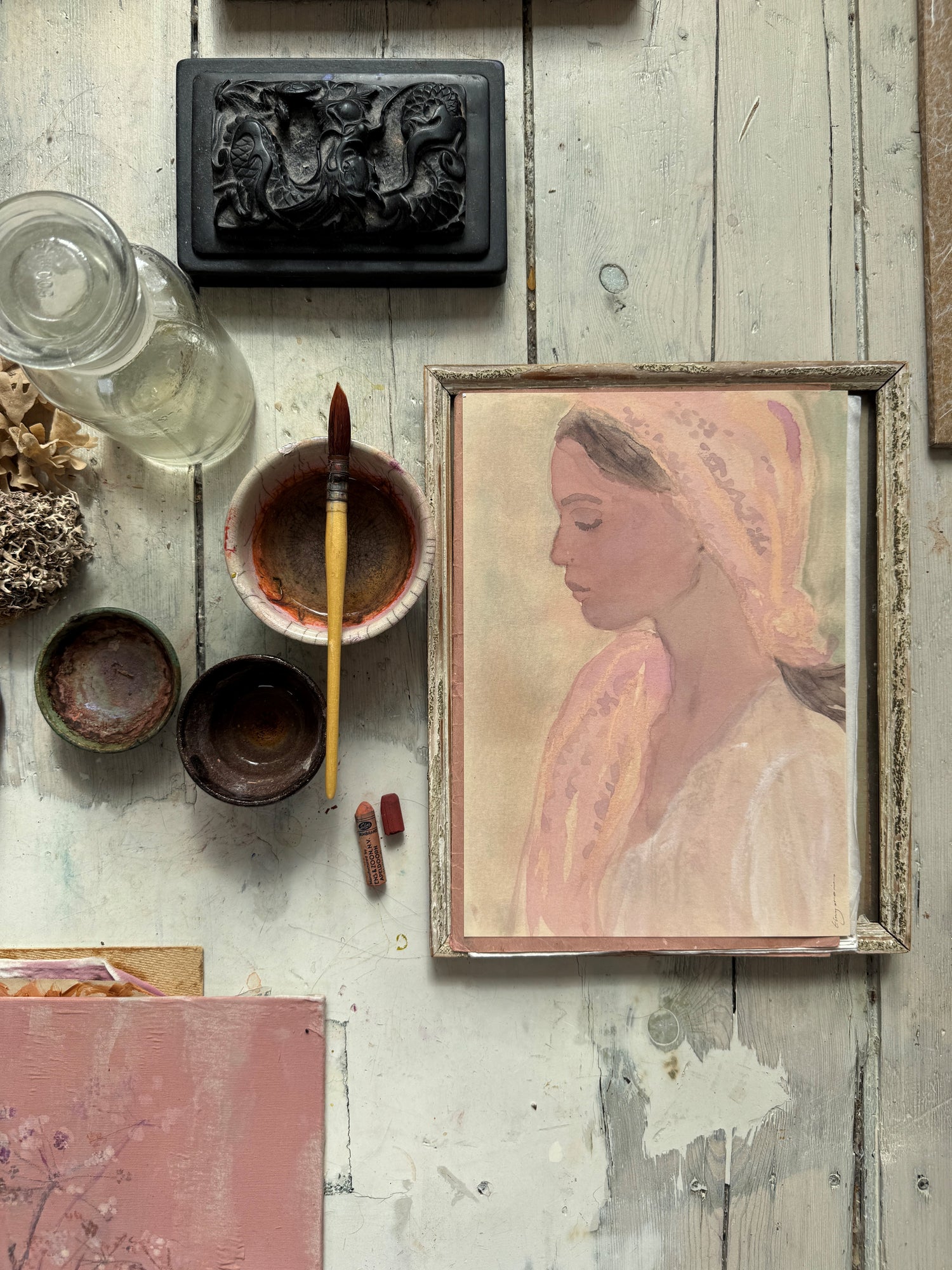 Framed watercolor portrait of a woman in profile wearing a pink headscarf, surrounded by paintbrush, jars, and art supplies
