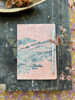 notebook in size DIN A5 showing an abstract landscape in soft colours with a pencil laying beside it