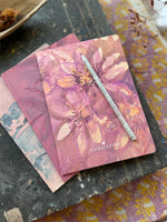 Three notebook laying on a wooden table. The notebook laying on top features a pink passion flower aquarell drawing.