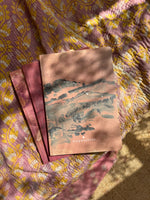 Three notebook laying on colorful fabric. The notebook laying on top features a drawing of a mountainside lanscape.