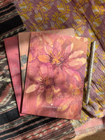 Three notebook laying on colorful fabric. The notebook laying on top features a pink passion flower aquarell drawing.