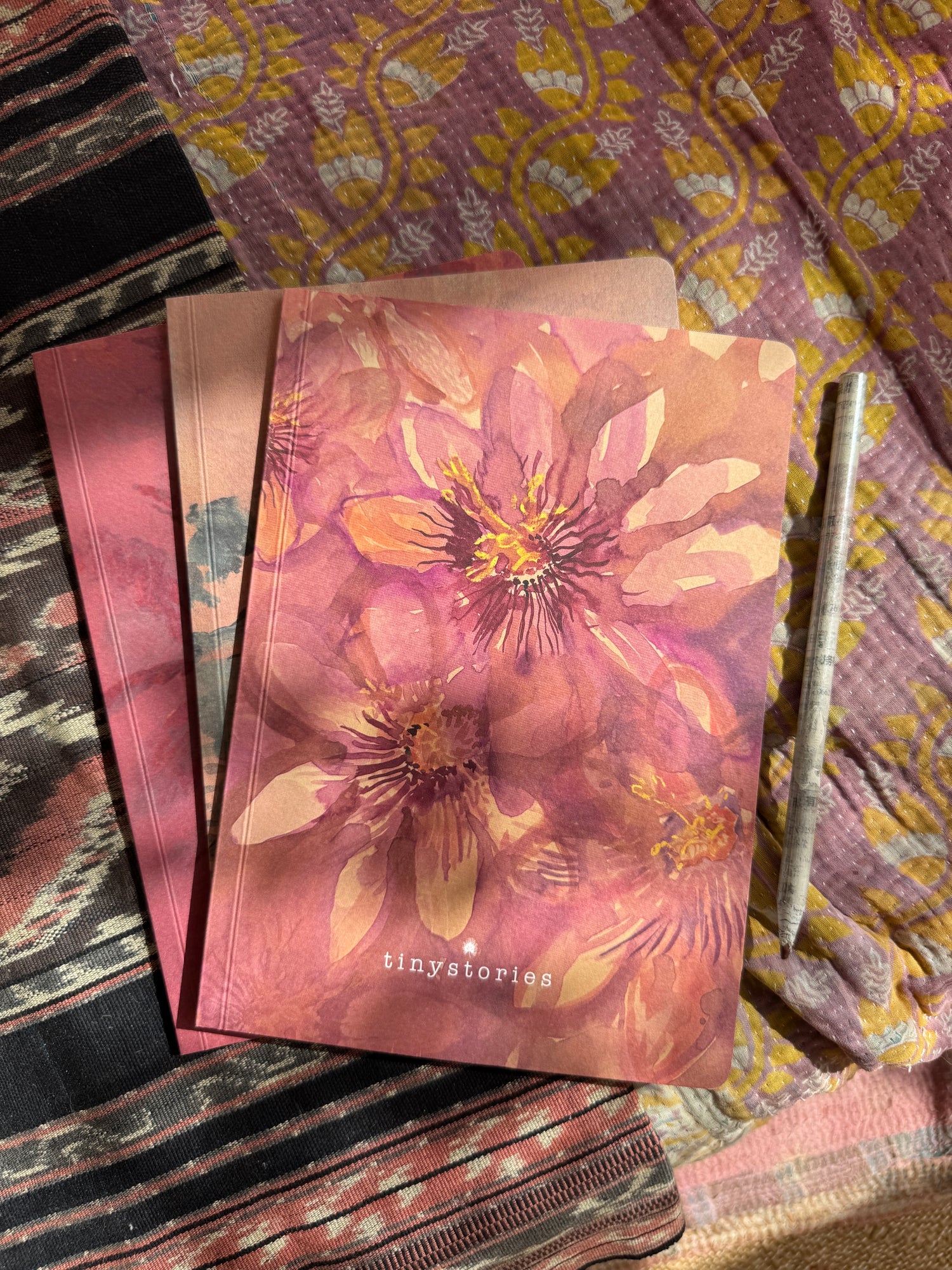Three notebook laying on colorful fabric. The notebook laying on top features a pink passion flower aquarell drawing.