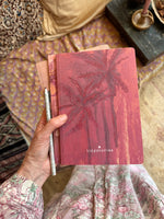 A hand holding three notebooks. The notebook laying on top features a drawing in shades of pink of palmtrees.