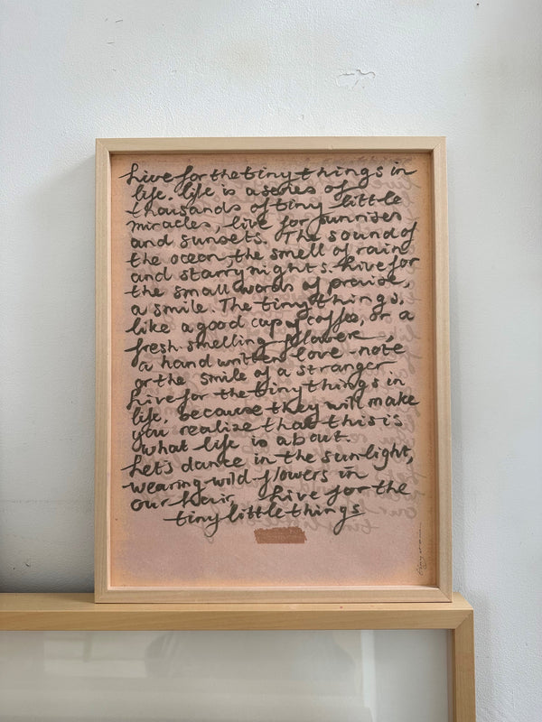 Close-up of framed handwritten poetic text on pink paper with artistic shadow play on rustic wooden floor

